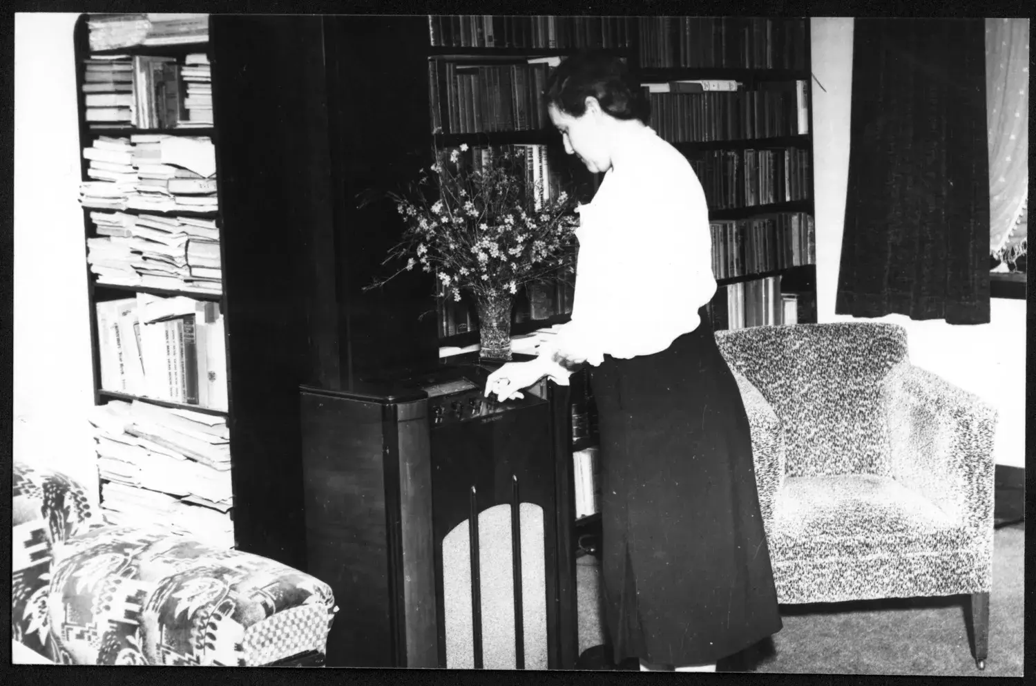 Elsie Curtin standing in a living room with a bookshelf, a lounge chair and a side table with a crystal vase with flowers.  
