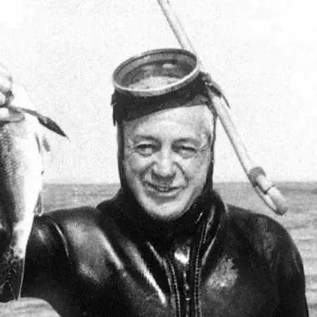 Harold Holt coming out of the ocean holding a fish in his hand, wearing a wetsuit with a snorkel mask on his head. 