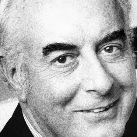 Headshot of Gough Whitlam smiling at the camera with his hand up.