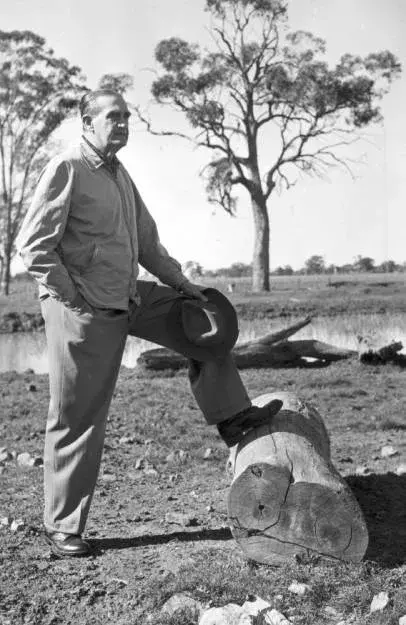 John McEwen is standing with one foot resting on a large fallen log. He holds a hat in his hands and looks out over his farm. 