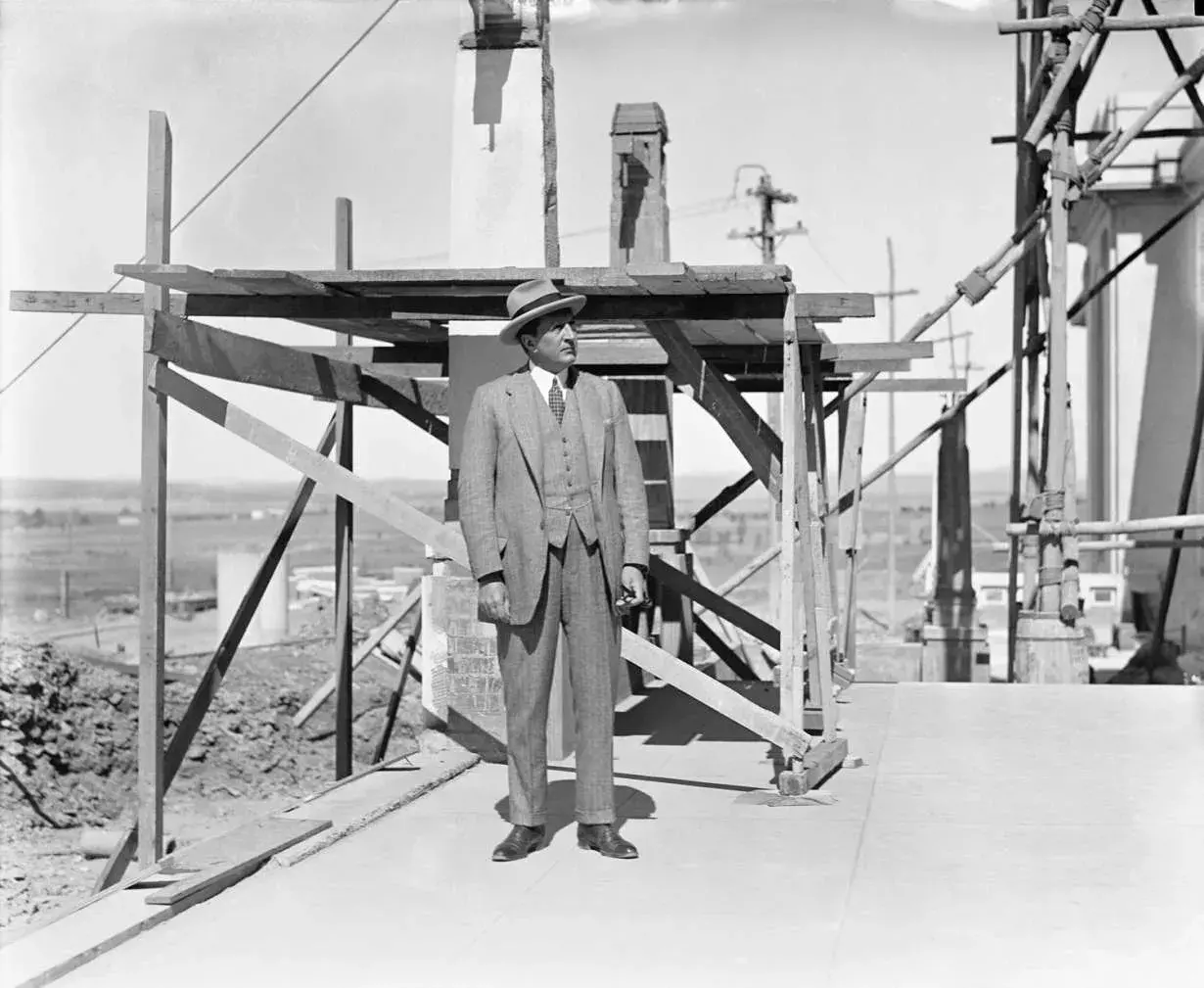 Prime Minister Stanley Bruce is dressed in a 3 piece suit and a hat, standing on the construction site of Old Parliament House with scaffolding behind him.  