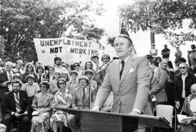 Malcolm Fraser stands at a lectern giving a speech with people surrounding him.  