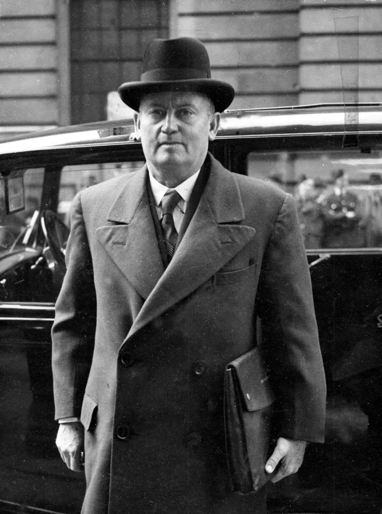 Frank Forde wearing a hat and coat and holding a leather satchel under his arm as he walks into a building. 