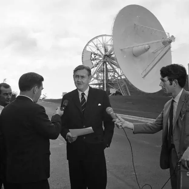 John Gorton stands in front of a satellite dish and is interviewed by a reporter. 