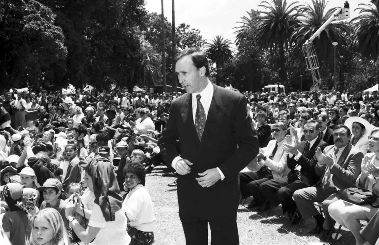 Paul Keating is dressed in a suit and surrounded by seated people outdoors listening to him.