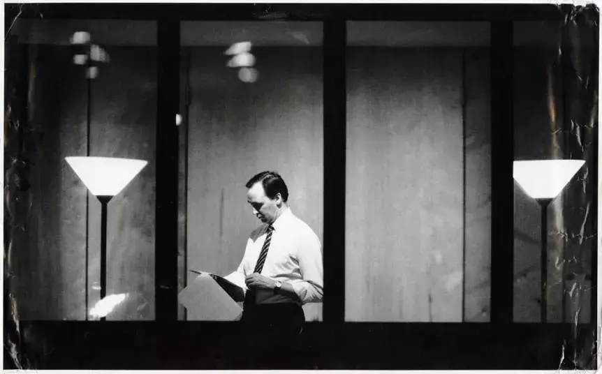 Paul Keating is in his office and reading a paper. He is viewed from outside looking through a window.  