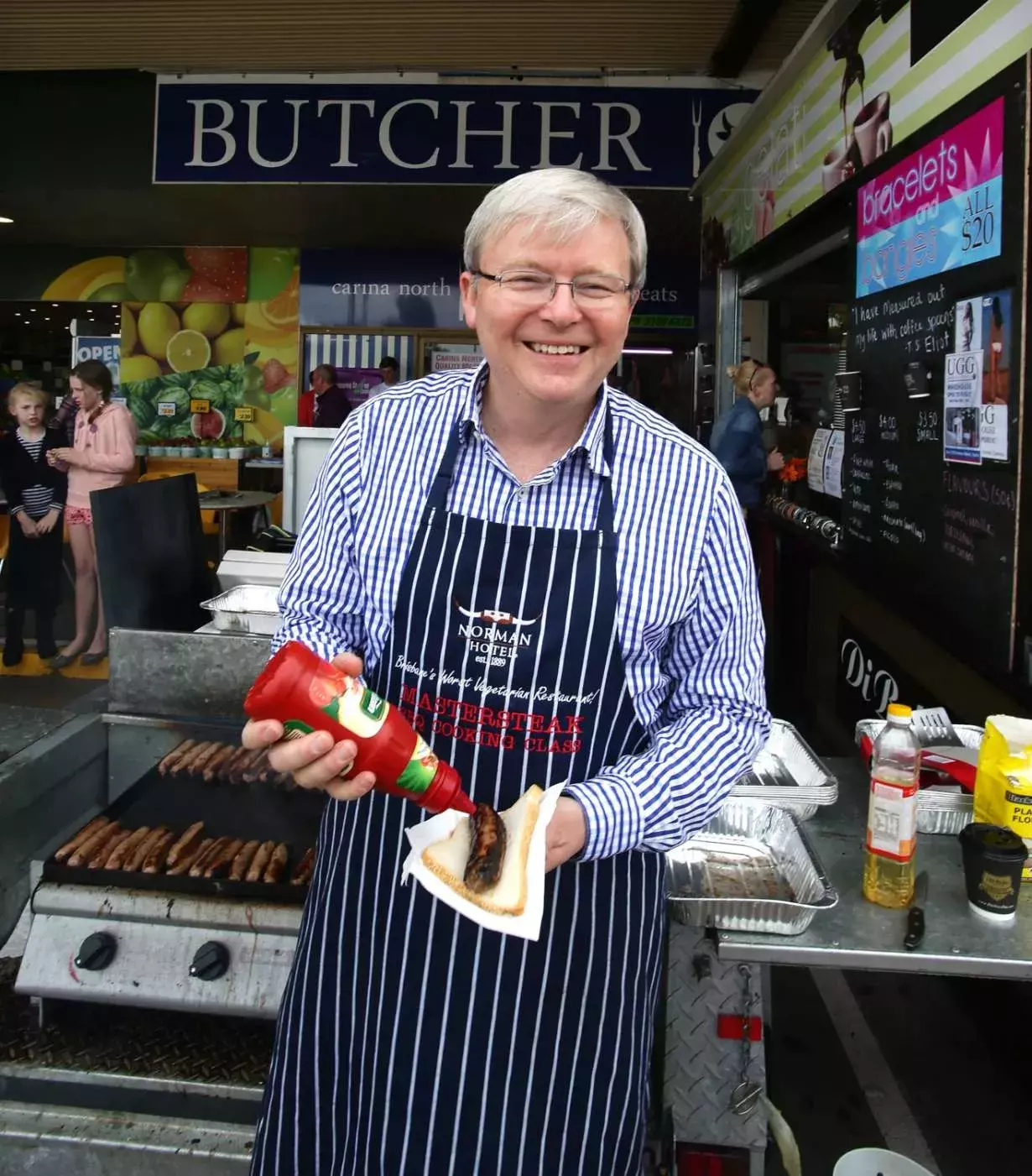 Kevin Rudd smiles at the camera while putting tomato sauce on a sausage at a sausage sizzle.  