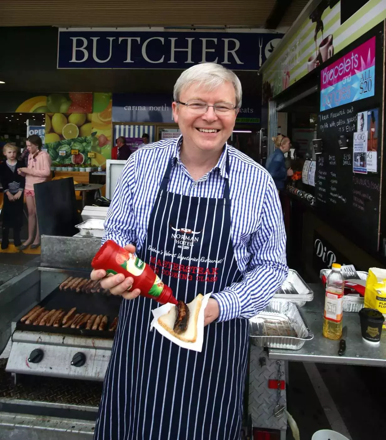 Kevin Rudd smiles at the camera while putting tomato sauce on a sausage at a sausage sizzle.