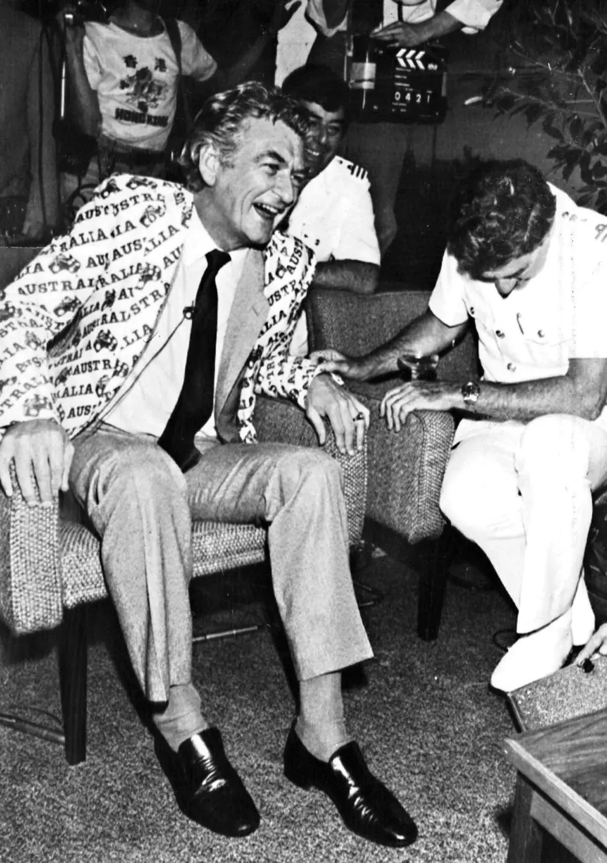 Bob Hawke sits in an armchair laughing with three men in uniform. He is wearing a suit jacket covered in the words 'Australia'.