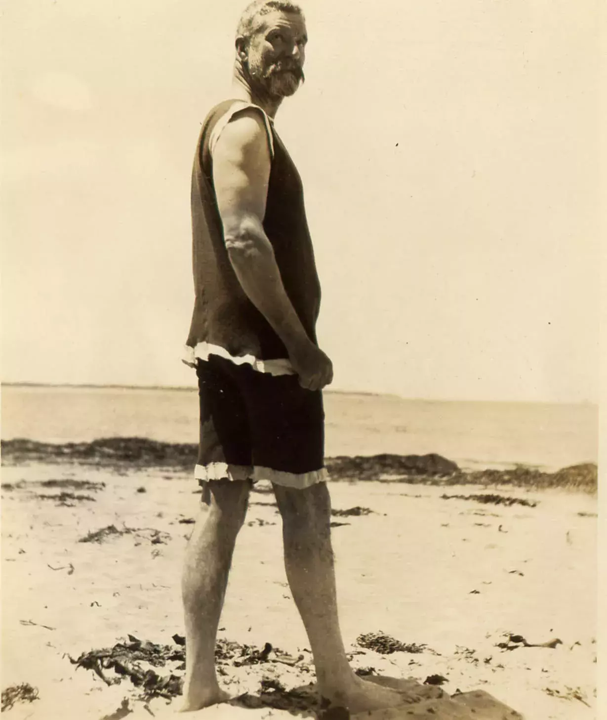 Alfred Deakin wearing a singlet and bathing shorts standing on the beach looking back at the camera.  