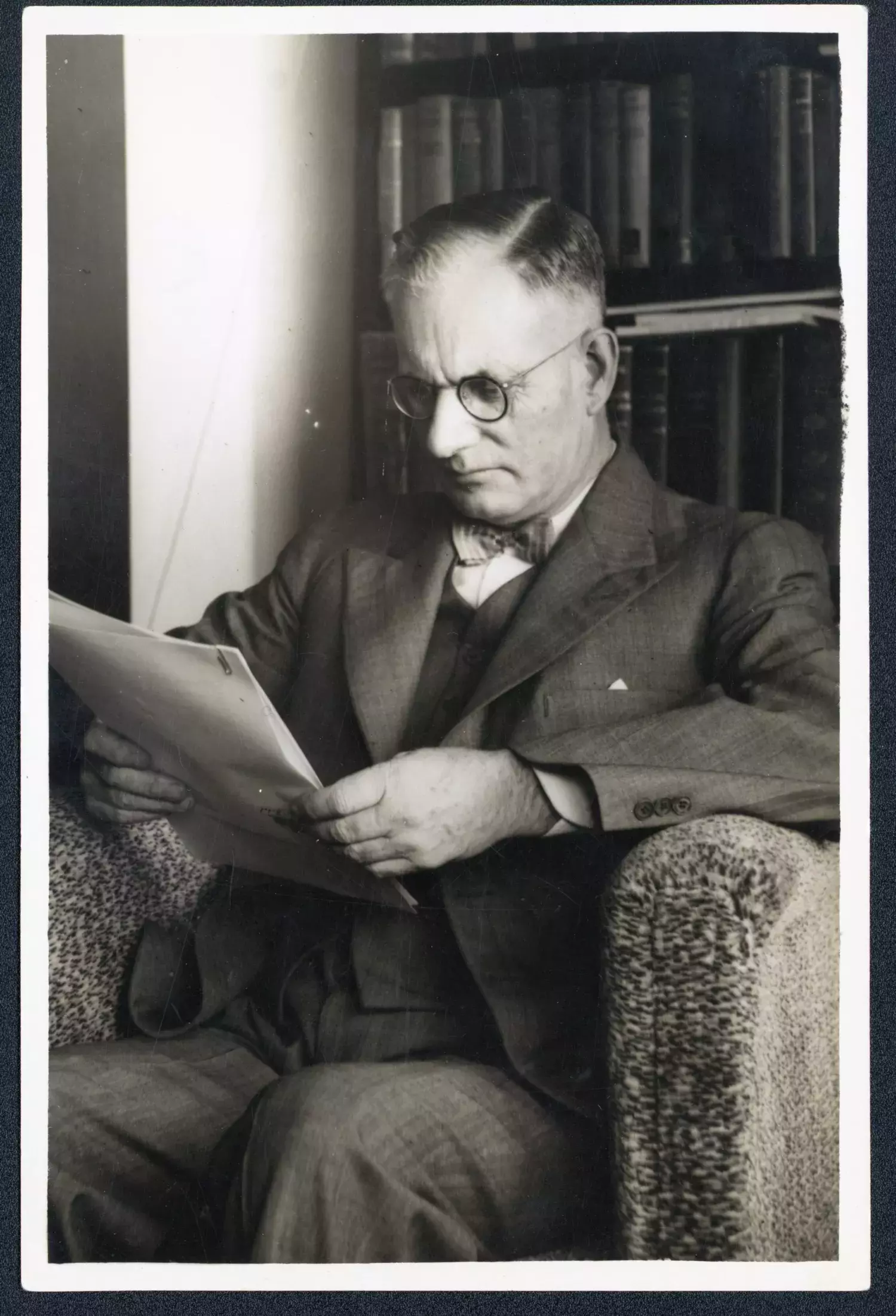 A black and white image of John Curtin reading in an armchair with bookcases behind hiim. 