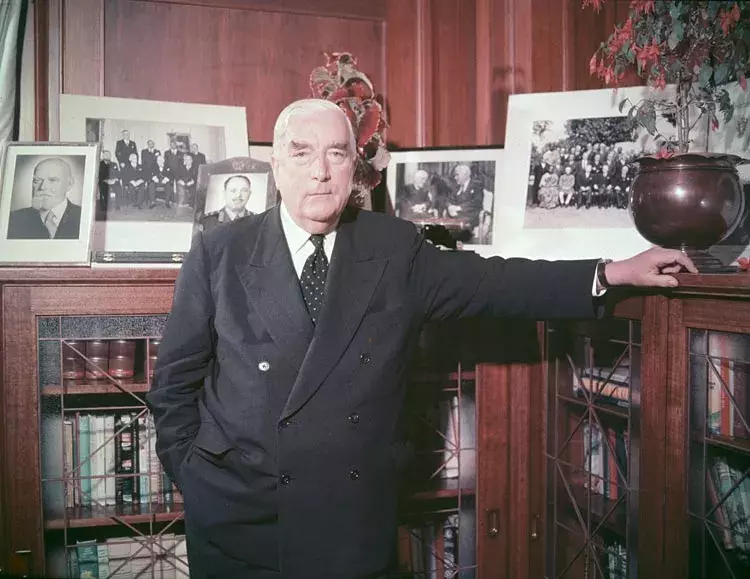 Robert Menzies leans on a bookshelf that has a collection of photos. 